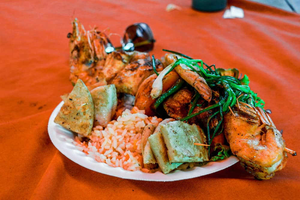 White plate on a red tablecloth with rice, vegetables and a skewer of different seafood.