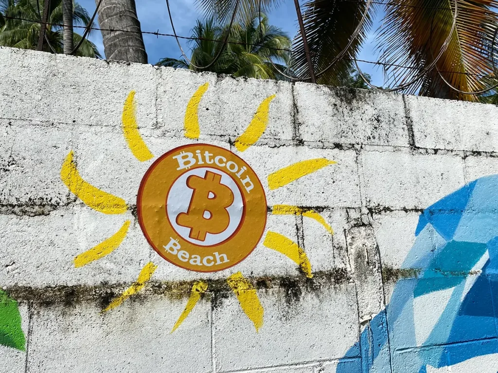 White brick wall with blue paint on one edge. Orange logo of "Bitcoin Beach" in the centre.