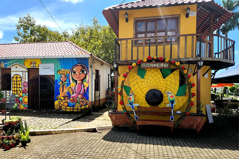 Yellow building with a ring of flowers and birds on it. Beside it, another building painted with indigenous art from El Salvador.